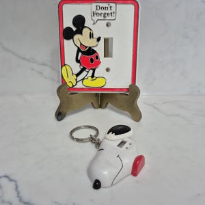 May include: A white light switch cover with a red border and a cartoon image of Mickey Mouse. The text "Don't Forget!" is in a speech bubble. A Snoopy keychain is in the foreground.
