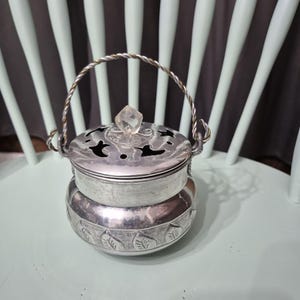 May include: A silver metal container with a lid and handle. The container has a decorative floral pattern around the base and a clear crystal-like knob on top. The handle is twisted silver wire.