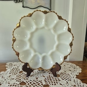 May include: A round white milk glass egg plate featuring twelve oval indentations arranged in a circle around a central depression. The plate has a scalloped edge trimmed with a metallic gold finish and is supported by a dark brown wooden stand.