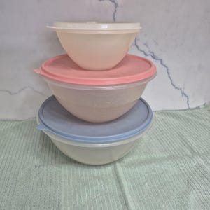 May include: Three plastic mixing bowls with lids stacked on top of each other. The top bowl has a white lid, the middle bowl has a pink lid, and the bottom bowl has a blue lid. The bowls are sitting on a green cloth.