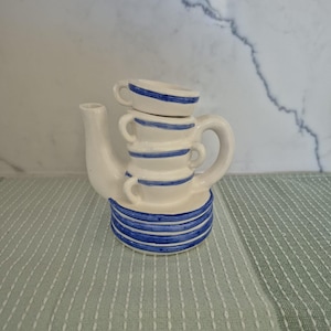 May include: A white ceramic teapot with blue horizontal stripes and a stack of three small teacups. The teapot has a curved handle and a spout. The teacups are also white with blue stripes. The teapot is sitting on a light green and white striped surface.