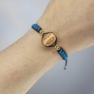 May include: A handmade bracelet featuring a light-colored wooden bead with etched designs. The bracelet has black and blue braided cord with gold-colored accents. The bracelet is adjustable.