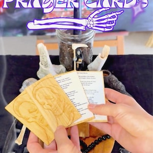 May include: Wooden prayer cards with skull carvings and text reading "DIOSA SANTA MUERTE PRAYER CARDS." The image includes a jar, a small figurine, and the text "I OFFER TO YOU: SANTA MUERTE PRAYER CARDS."