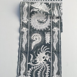 May include: Dark teal scarf with white detailed patterns. The scarf features a repeating design of paisley, fish, and bird motifs. The edges have a fringe detail. The scarf is folded and displayed against a light gray background.