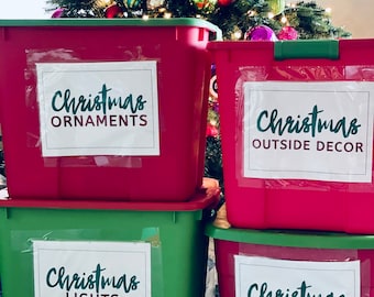 May include: Four red and green plastic storage bins with white labels that say "Christmas Ornaments", "Christmas Outside Decor", "Christmas Lights", and "Christmas Garlands".