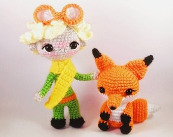 May include: A crocheted toy of The Little Prince wearing a yellow scarf and a fox with orange fur. The Little Prince is wearing a green outfit and has a blonde wig with orange goggles. The fox is sitting with its tail curled around its body.
