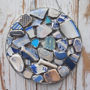 May include: A round, mosaic-style wall hanging composed of various blue and white ceramic shards. The pieces are arranged within a silver metal frame and suspended by a chain. The ceramic fragments feature floral and geometric patterns.