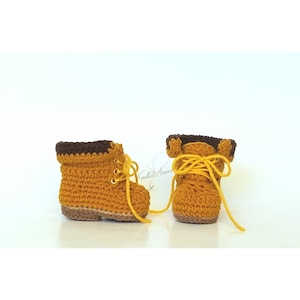 May include: A pair of crocheted baby booties in a mustard yellow color with brown trim and yellow laces.