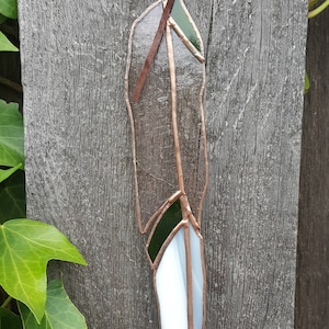 May include: A stained glass feather with a copper frame and a brown leather cord for hanging. The feather features white, clear, and green glass pieces. The background is a weathered wooden surface with green ivy.