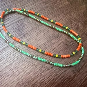 Könnte beinhalten: Eine zweireihige Perlenkette aus kleinen, runden Glasperlen in leuchtendem Neonorange, hellem Limettengrün und Gelb, durchsetzt mit metallisch bronzefarbenen Perlen, die in einem sich wiederholenden kreisförmigen Muster angeordnet sind.