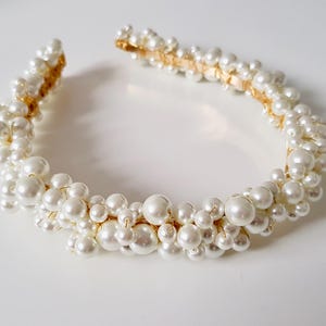 May include: A white pearl headband with a gold-colored band. The headband is adorned with numerous white pearls of varying sizes, clustered together to create a full, textured look. The pearls are arranged around the entire band.