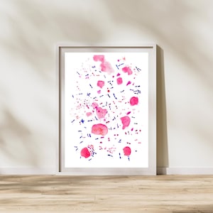 May include: A framed abstract watercolor print featuring pink and blue splatters and shapes on a white background. The artwork is displayed in a white frame and leans against a white wall, with a wooden floor in the foreground.