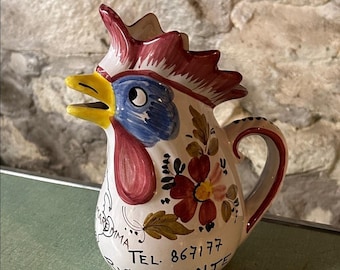 Puede incluir: Jarra de cerámica con forma de gallo, con cresta roja, pico amarillo y cara azul y blanca. La jarra está decorada con motivos florales y el texto "TEL. 867177 RISTORANTE". El asa es roja.