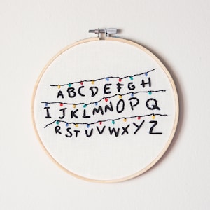 May include: Embroidered hoop art featuring the alphabet in black lettering with colorful string lights. The letters are arranged in three rows, with the first row reading "ABCDEFGH", the second row reading "IJKLMNOPQ", and the third row reading "RSTUVWXYZ".