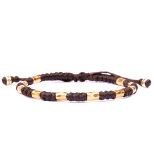 May include: A dark brown cord bracelet featuring a series of cylindrical, hammered gold-tone metal beads spaced evenly along the band. The bracelet includes adjustable knotted ends with small, engraved gold-tone metal caps at the tips.