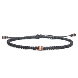 May include: A black braided cord bracelet with a copper accent. The bracelet is tied with a slip knot.