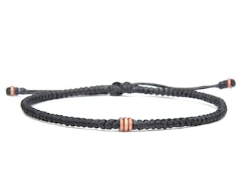 May include: A black cord bracelet featuring a braided design with three small copper-colored metal rings centered on the band. The bracelet includes an adjustable sliding knot closure with two additional copper-colored beads at the ends of the cords.