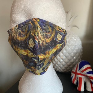 May include: A face mask with a colorful, abstract design featuring a painting of houses. The mask is made of fabric and has a black elastic band.
