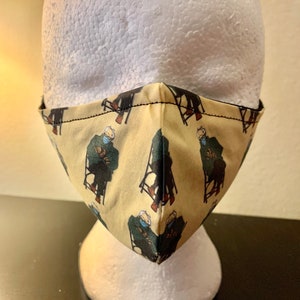 May include: A beige face mask with a repeating pattern of a person sitting in a chair. The person is wearing a green jacket and a blue hat.