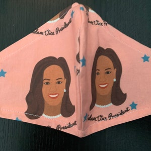 May include: A pink face mask with a repeating pattern of a woman's face and the text "Madam Vice President" and blue stars.