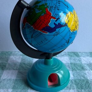 Pode incluir: Um pequeno globo com oceanos azuis e continentes multicoloridos, montado num arco meridiano de plástico preto. A base é um afia-lápis de plástico azul-petróleo com uma abertura interior rosa, exibindo texto impresso de países e oceanos.