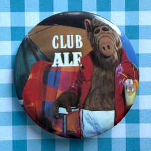 May include: A circular pinback button featuring a brown, furry creature wearing a red jacket and white shorts. The creature holds a drink with a small umbrella. The text CLUB ALF is printed in white letters above the creature against a background.