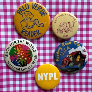May include: Five circular pinback buttons. One yellow button reads Palo Verde Reader with a cartoon character. One cream button shows a train and Reading Railroad 1985. One white button features colorful hands and Reach for the World National Library Week. One blue button shows a building and Reading Road Trip U.S.A. One yellow button says NYPL.