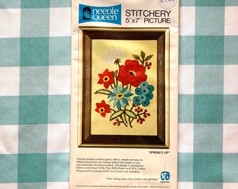 May include: Needlepoint embroidery kit featuring a floral design with red, blue, and white flowers. The kit includes colourful yarns, fabric, a needle, and instructions. The design is stamped on a 5x7 inch piece of fabric. The kit is titled "Stitchery 5"x7" Picture" and "Spring's Up".
