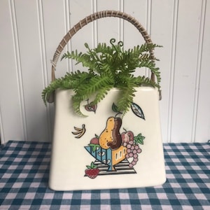 May include: White ceramic planter shaped like a handbag with a woven handle. The planter features a colorful illustration of fruit in a bowl. A green fern plant is growing inside the planter.