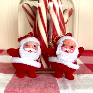 Pode incluir: Dois enfeites de Papai Noel de feltro vermelho com detalhes brancos e rostos pintados. Os enfeites estão pendurados em varinhas de doces em um frasco de vidro.