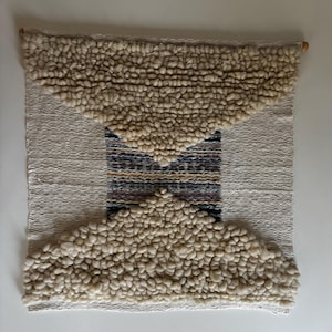 Puede incluir: Un tapiz de pared tejido beige y blanco con un diseño geométrico. El tapiz presenta una sección central de tejido tejido con rayas en tonos de azul, amarillo y marrón. Las secciones superior e inferior están hechas de hilo blanco esponjoso.