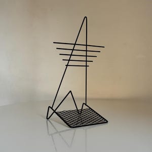 May include: A black metal sculpture with an abstract design. The sculpture features a triangular base with a grid pattern, a vertical line, and horizontal lines arranged in a tiered fashion. The sculpture is on a white surface.