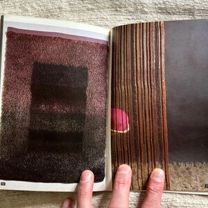 May include: A double-page spread from a magazine featuring abstract textile art. The left page displays a textured, rectangular design in shades of burgundy and black. The right page shows vertical, woven textures in brown and tan, with a pink accent.