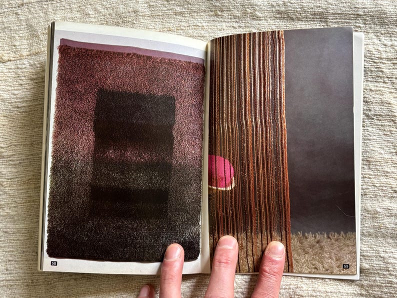 May include: A double-page spread from a magazine featuring abstract textile art. The left page displays a textured, rectangular design in shades of burgundy and black. The right page shows vertical, woven textures in brown and tan, with a pink accent.