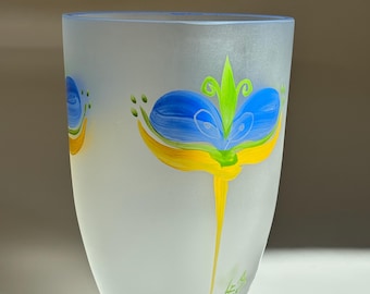 Puede incluir: Jarrón de vidrio esmerilado con diseño floral pintado a mano. Las flores son azules, amarillas y verdes. El jarrón tiene forma cilíndrica y un borde ligeramente ensanchado. El diseño se repite.