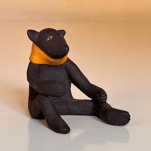 Puede incluir: Figura de oso negro con bufanda naranja, sentado con las piernas extendidas. El oso tiene orejas pequeñas y descansa sobre una superficie blanca. La figura es un artículo artesanal.