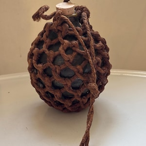 May include: A spherical dark green glass bottle encased in a thick brown knotted rope netting. The bottle features a light-coloured cork stopper at the top, with loose rope ends extending from the netting to form a handle or hanging loop.