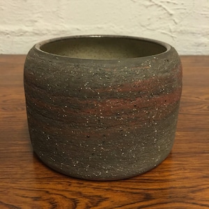 May include: A dark gray ceramic planter with a rough, textured surface and subtle red streaks. The planter has a rounded shape and a wide opening.