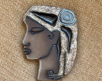May include: A ceramic wall hanging in the shape of a woman's profile. The piece features a dark brown face with closed eyes, black outlines, and a light brown textured hair design. A blue-green spiral adorns the hair. The overall color palette is earthy.