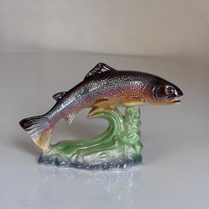 Könnte beinhalten: Eine Fischfigur aus Keramik mit einem dunkelgrauen und schillernd violetten Körper sowie einem hellen Bauch. Der Fisch ist in einer Sprungbewegung dargestellt und wird von einem grünen Sockel gestützt, der stilisierte Wasserwellen und Felsen zeigt.