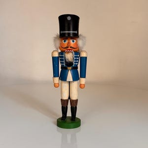 Peut inclure: Une figurine de casse-noisette en bois traditionnelle avec un uniforme bleu et blanc, un chapeau noir et une moustache et une barbe blanches. Le casse-noisette est debout sur une base verte.