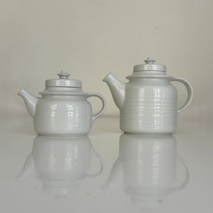 May include: Two white ceramic teapots, one larger than the other, with a textured surface. Both teapots have a lid and a spout.