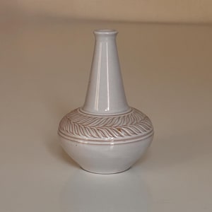 May include: A white ceramic vase with a slender neck and a rounded base. The base has a detailed leaf pattern in a lighter shade of white. The vase is a decorative item, suitable for displaying flowers or as a standalone accent piece.