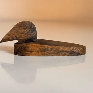 Op de afbeelding: Een donkerbruin houten eendenbeeldje met een gegraveerd hoofd en een glad, rond lichaam. De eend zit op een wit oppervlak.