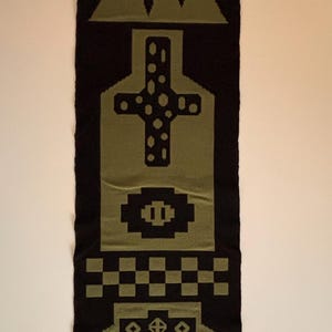 Puede incluir: Textil tejido en negro y verde oliva con patrones geométricos. El diseño incluye una cruz, formas de diamante y líneas verticales. El textil es rectangular y parece ser una pieza decorativa.