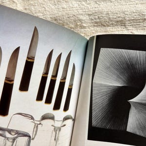 May include: An open book displays a collection of knives with dark brown handles and silver blades. The knives are arranged on a light blue background. The opposite page features a black and white abstract design.