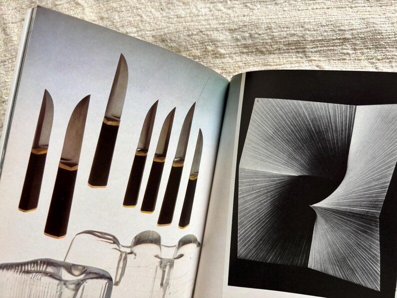 May include: An open book displays a collection of knives with dark brown handles and silver blades. The knives are arranged on a light blue background. The opposite page features a black and white abstract design.