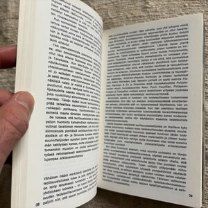 May include: An open book with text in a foreign language, held by a hand. The pages are white with black text. The book is open to pages 38 and 39. The background is a textured, neutral surface.