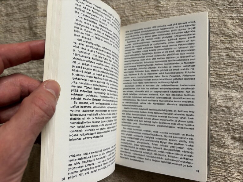 May include: An open book with text in a foreign language, held by a hand. The pages are white with black text. The book is open to pages 38 and 39. The background is a textured, neutral surface.