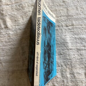 May include: A hardcover book titled "Suomen taideteollisuus" with a blue and black abstract design on the cover. The spine displays the title and author, Ulf H&aring;rd af Segerstad, in white text. The book is set against a neutral background.